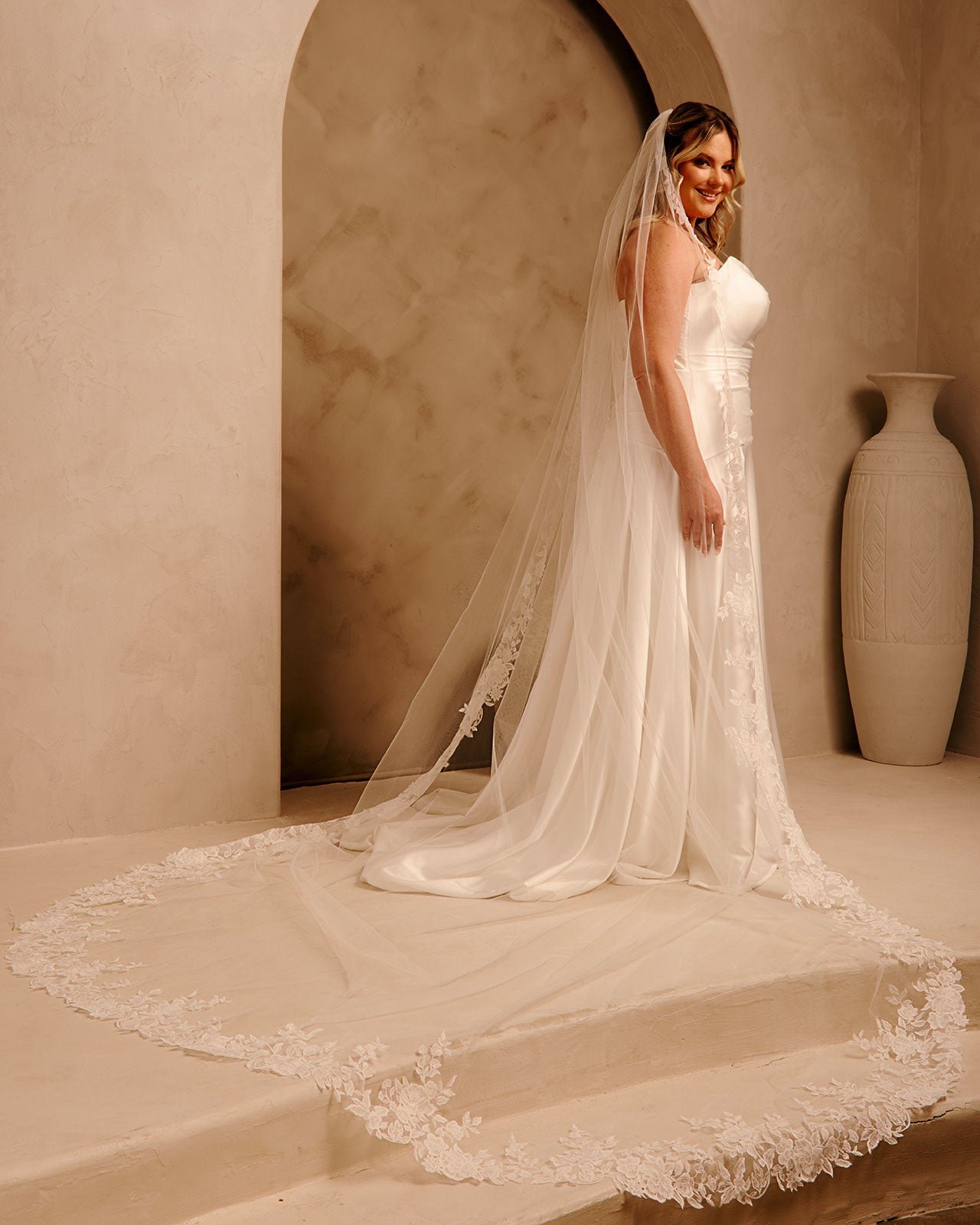 Rose Lace Royal Cathedral Bridal Veil Cassandra Lynne - A woman wearing a sleeveless wedding gown with a long sheer veil and lace-trimmed train, standing in a warm beige architectural setting with a large vase in the background.