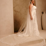 Rose Lace Royal Cathedral Bridal Veil Cassandra Lynne - A woman wearing a sleeveless wedding gown with a long sheer veil and floral lace trim, standing in a warm beige architectural setting with arches and a large vase.