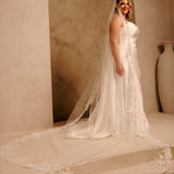 Rose Lace Royal Cathedral Bridal Veil Cassandra Lynne - A woman wearing a sleeveless wedding gown with a long sheer veil and floral lace trim, standing in a warm beige architectural setting with arches and a large vase.