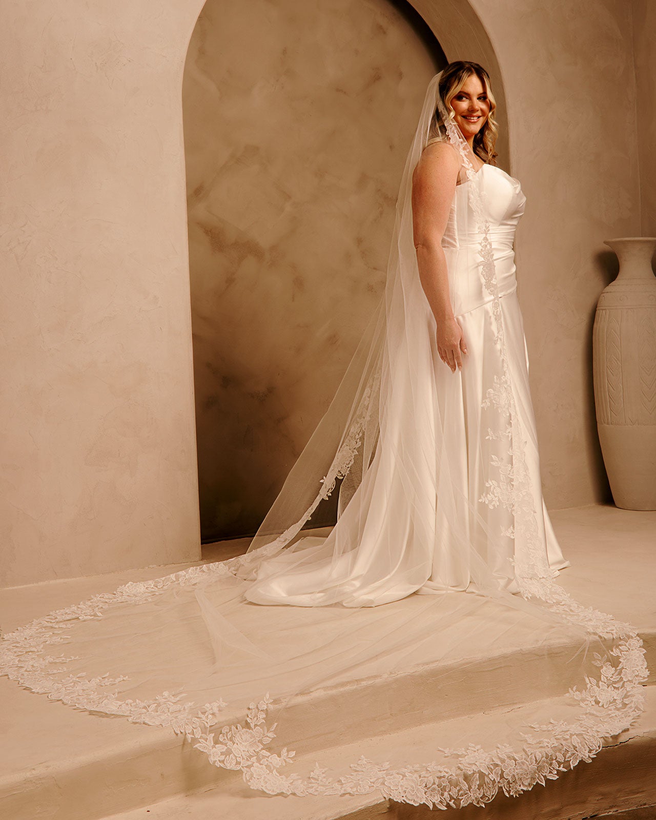 Rose Lace Royal Cathedral Bridal Veil Cassandra Lynne - A woman wearing a sleeveless wedding gown with a long sheer veil and floral lace trim, standing in a warm beige architectural setting with arches and a large vase.