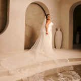 Royal Cathedral Veil with Blusher - Cassandra Lynne A woman wearing an ivory strapless wedding gown with a long sheer veil and a large lace-trimmed train stands on stone steps in a warm beige architectural setting with arches and large vases.