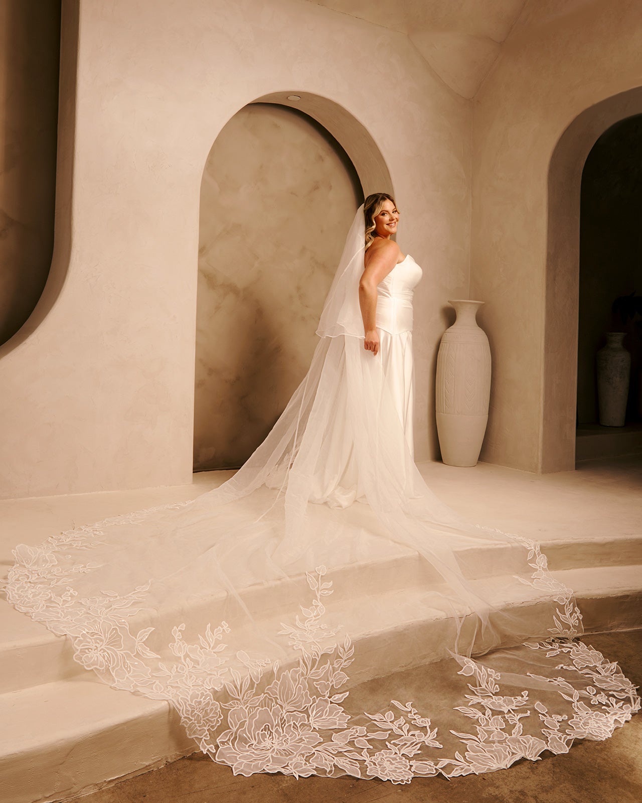 Royal Cathedral Veil with Blusher - Cassandra Lynne A woman wearing an ivory strapless wedding gown with a long sheer veil and a large lace-trimmed train stands on stone steps in a warm beige architectural setting with arches and large vases.