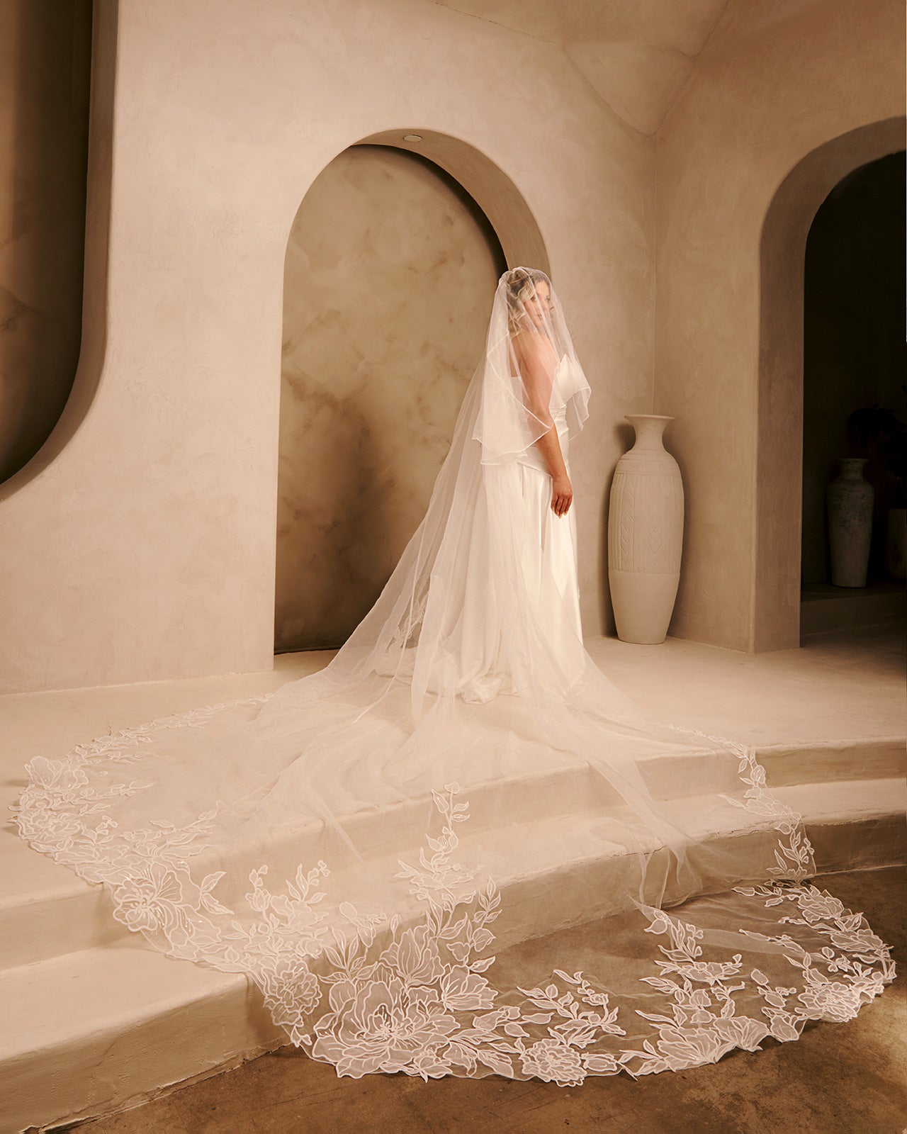Royal Cathedral Veil with Blusher - Cassandra Lynne A bride in an ivory wedding gown with a long lace-trimmed train and veil stands on stone steps in a warm, beige architectural setting with arches and large vases.