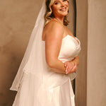 Royal Cathedral Veil with Blusher - Cassandra Lynne A woman wearing a strapless ivory wedding dress with a long layered veil, standing in a warm-toned indoor setting with a textured beige wall.