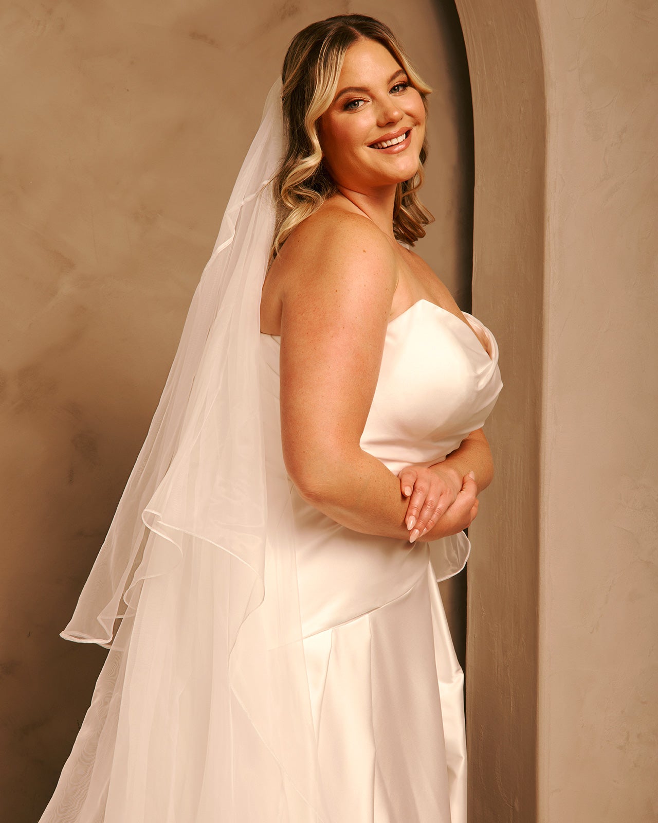 Royal Cathedral Veil with Blusher - Cassandra Lynne A woman wearing a strapless ivory wedding dress with a long layered veil, standing in a warm-toned indoor setting with a textured beige wall.