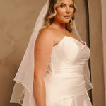 Royal Cathedral Veil with Blusher - Cassandra Lynne A woman wearing a strapless ivory wedding dress with a matching sheer veil, posed against a warm beige studio backdrop.