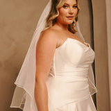 Royal Cathedral Veil with Blusher - Cassandra Lynne A woman wearing a strapless ivory wedding dress with a matching sheer veil, posed against a warm beige studio backdrop.