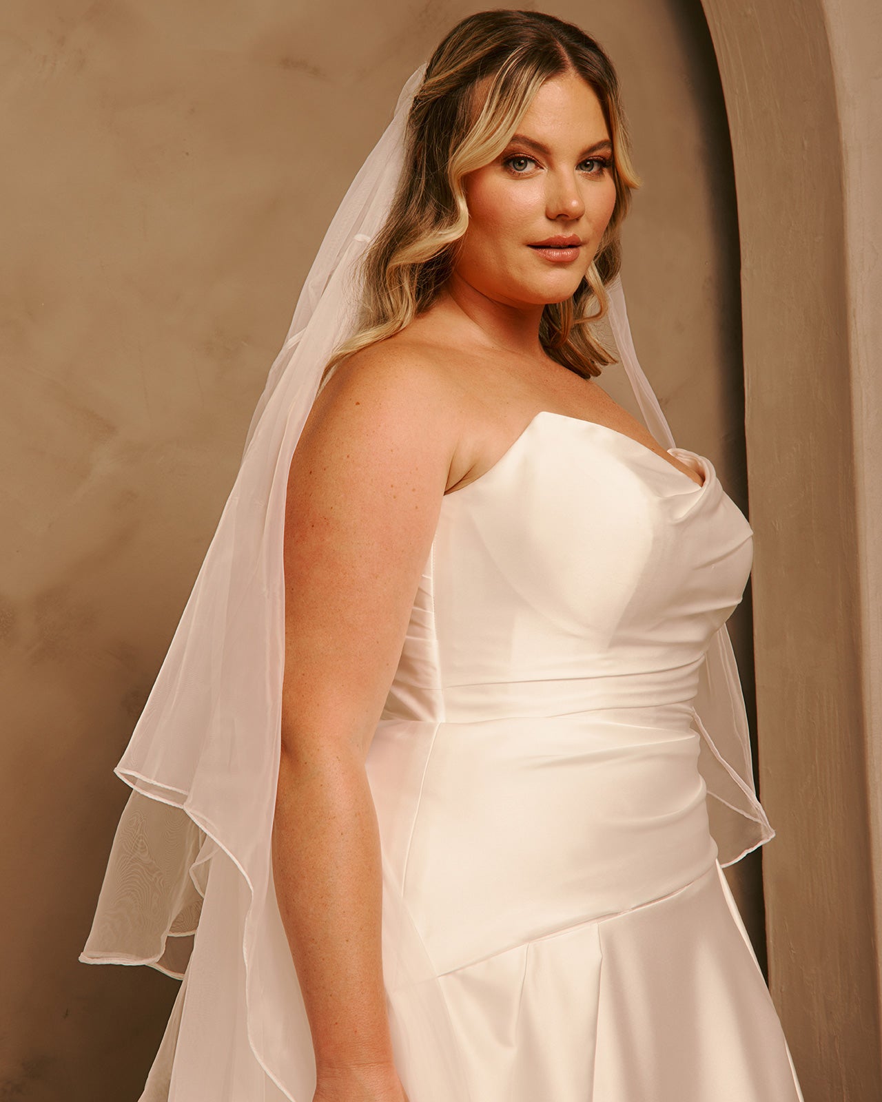 Royal Cathedral Veil with Blusher - Cassandra Lynne A woman wearing a strapless ivory wedding dress with a matching sheer veil, posed against a warm beige studio backdrop.