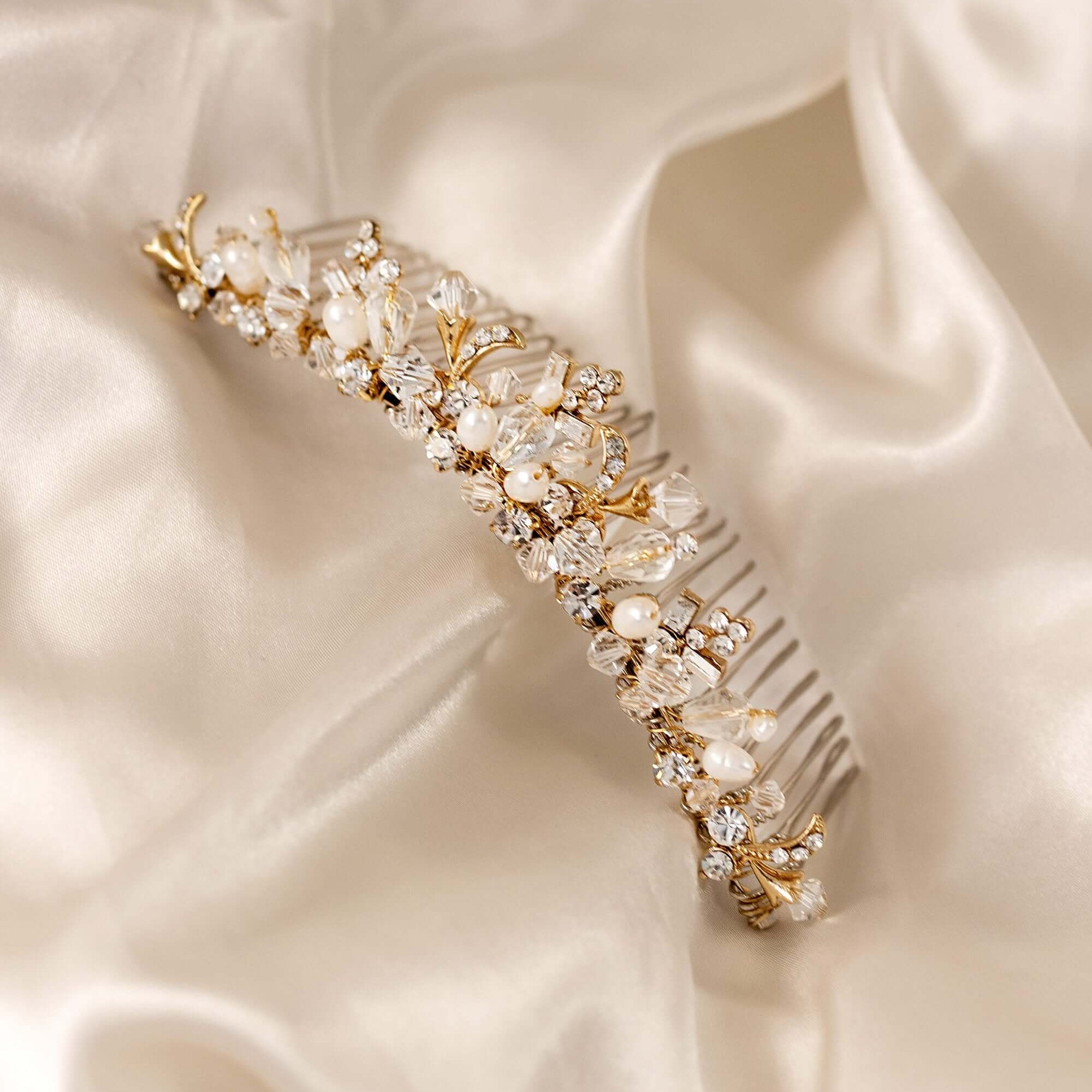 Gold Tiara Comb with Freshwater Pearls A gold bridal hair comb embellished with clear crystals, faux pearls, and delicate gold leaf details, displayed on a draped cream satin fabric.