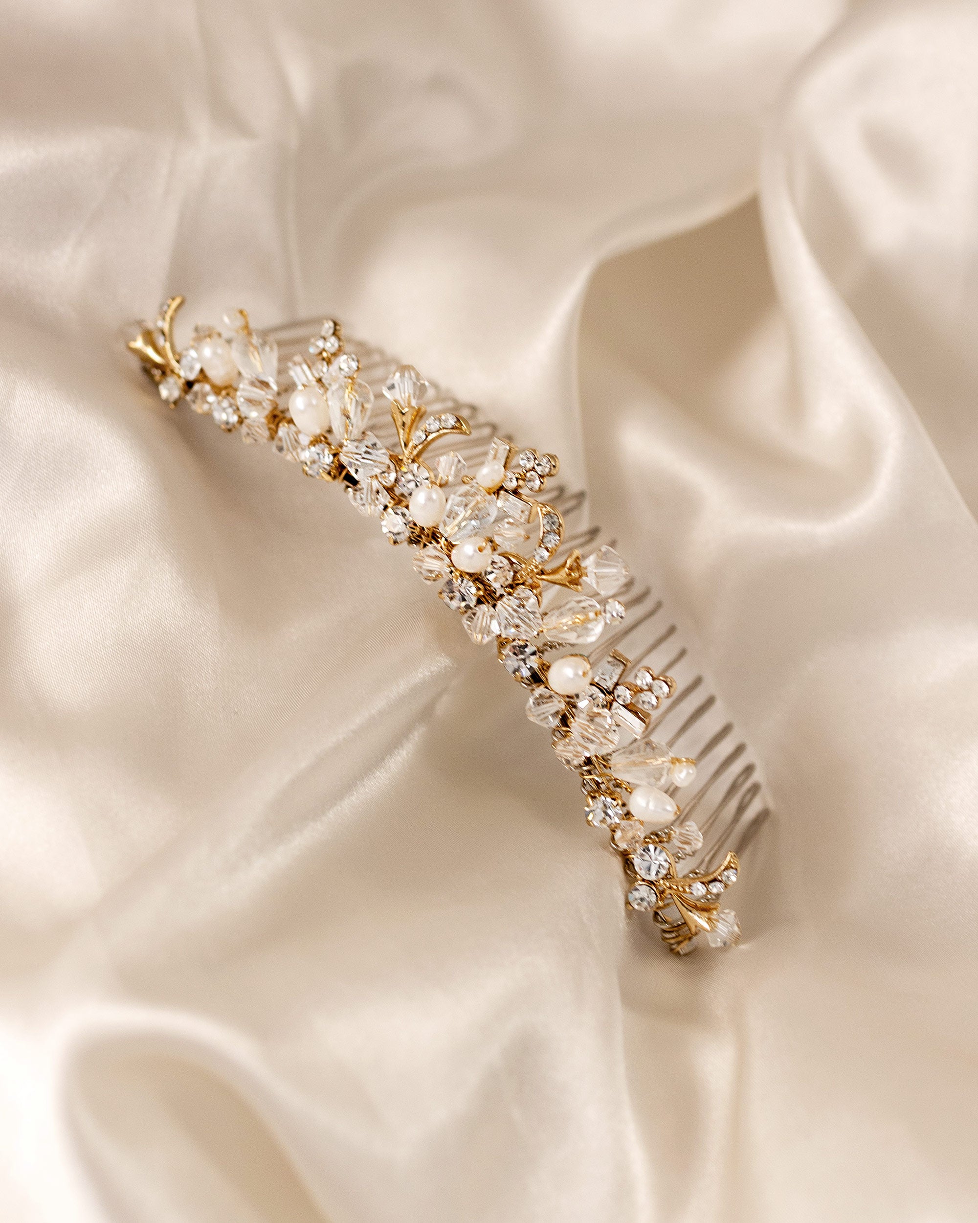 Gold Tiara Comb with Freshwater Pearls A gold bridal hair comb embellished with clear crystals, faux pearls, and delicate gold leaf details, displayed on a draped cream satin fabric.