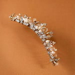 Gold Tiara Comb with Freshwater Pearls A delicate gold floral tiara adorned with clear crystals and white pearls, displayed on a warm brown background.