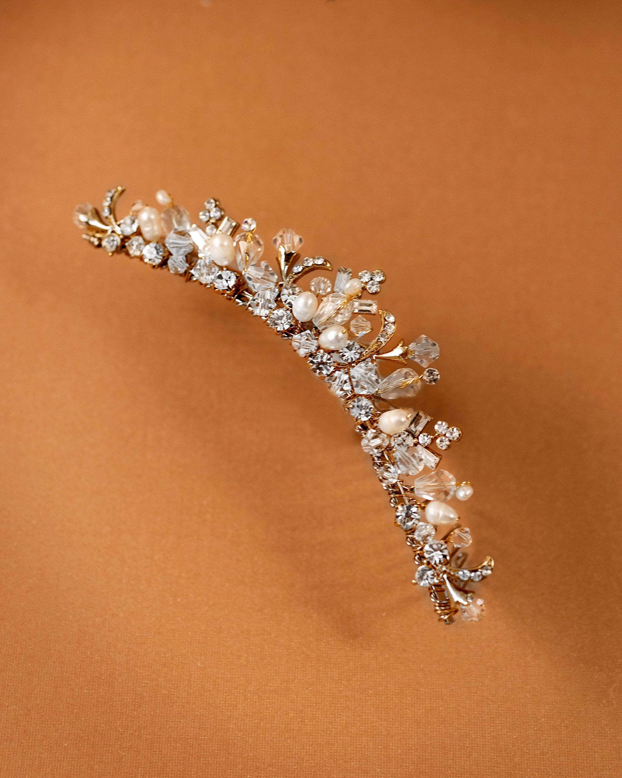 Gold Tiara Comb with Freshwater Pearls A delicate gold floral tiara adorned with clear crystals and white pearls, displayed on a warm brown background.