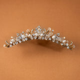 Gold Tiara Comb with Freshwater Pearls A gold floral crown headband embellished with clear crystals and white faux pearls, displayed against a solid warm brown background.