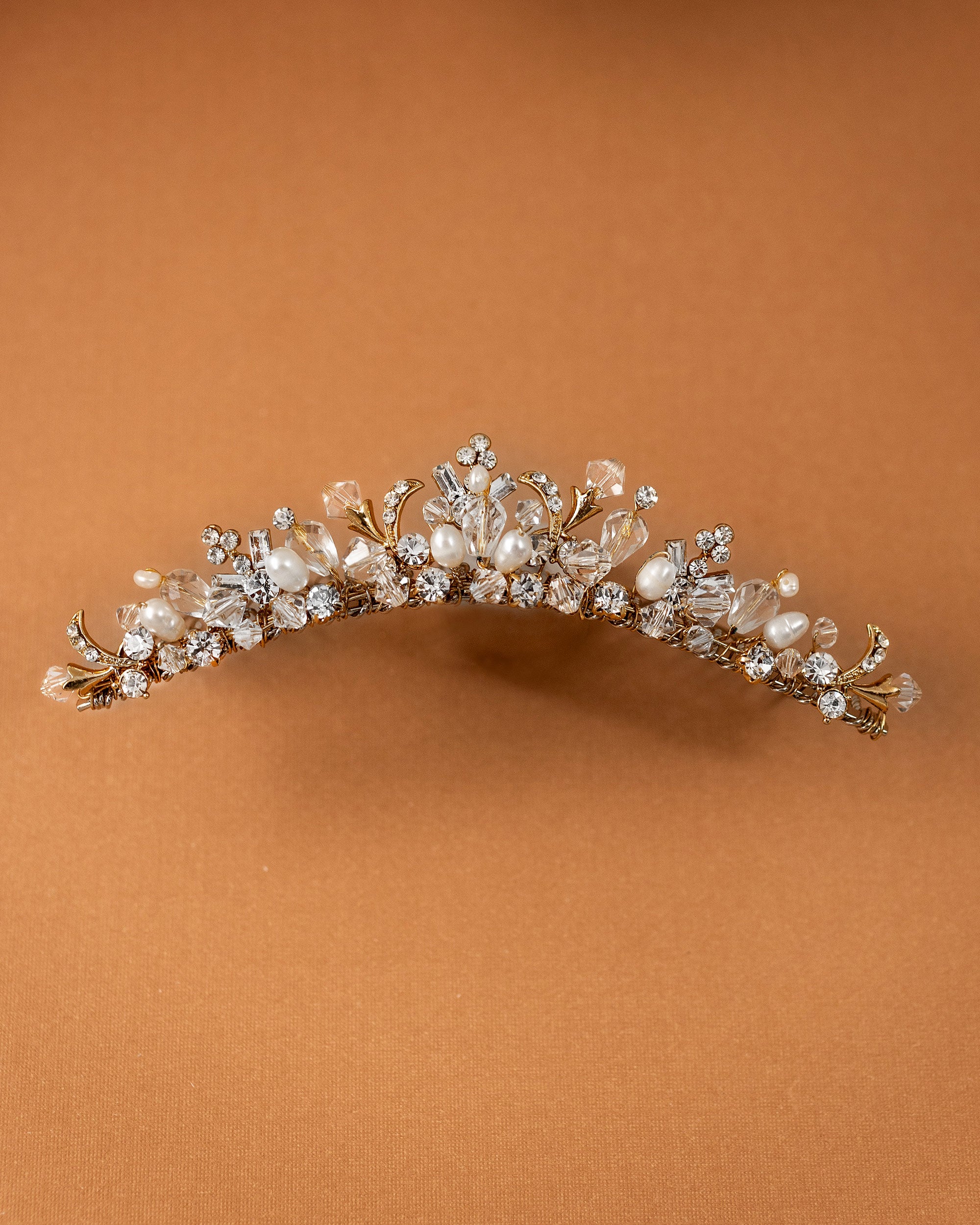Gold Tiara Comb with Freshwater Pearls A gold floral crown headband embellished with clear crystals and white faux pearls, displayed against a solid warm brown background.