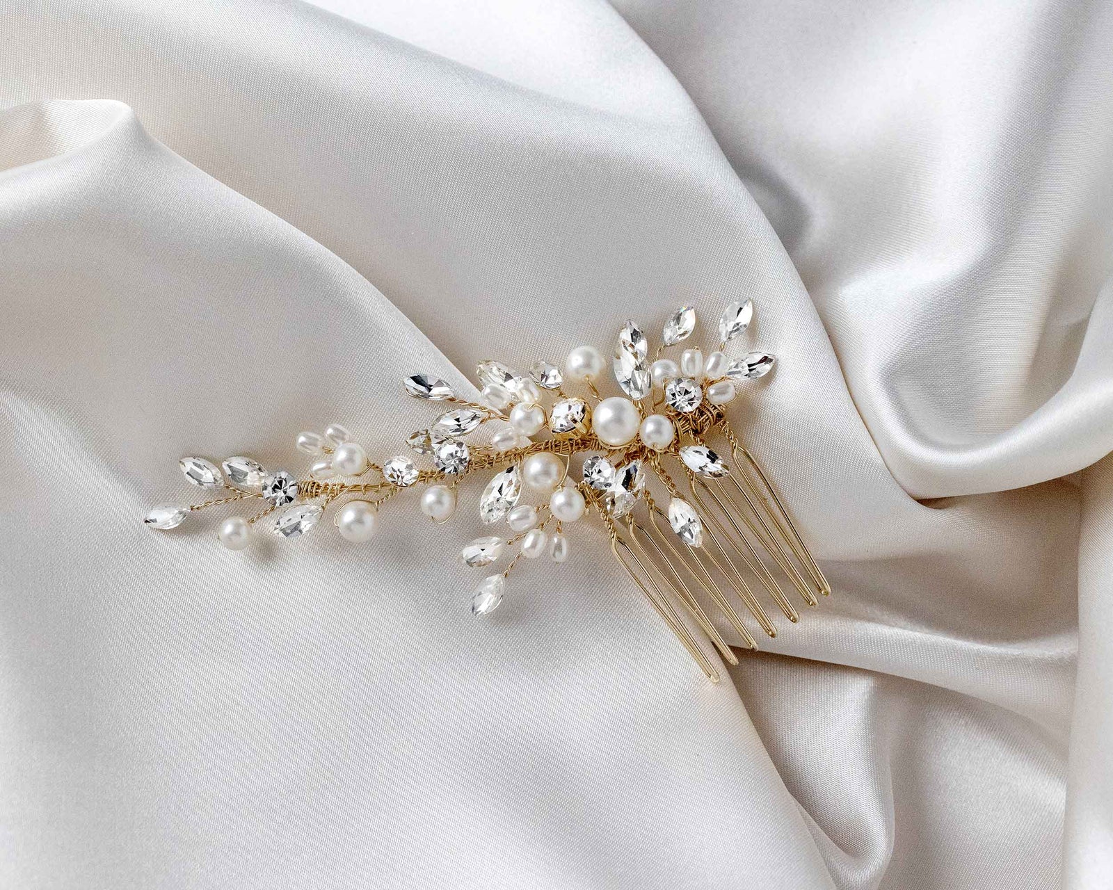 A silver-colored bridal hair comb featuring matte ivory pearls and marquise-cut rhinestones, placed on a grey background next to a piece of white lace fabric.