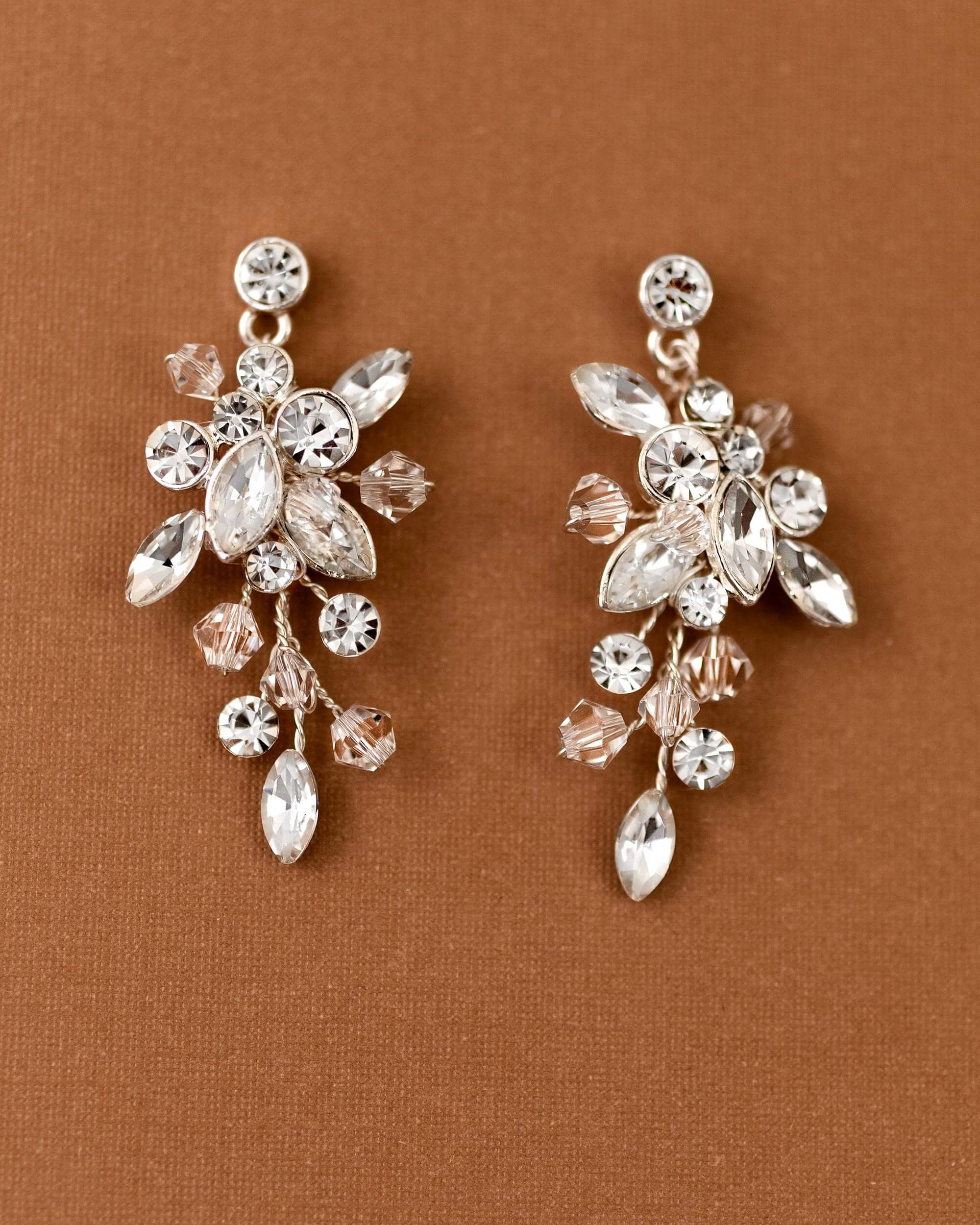 A pair of silver-toned floral drop earrings adorned with clear and pale pink rhinestones, displayed on a textured brown background.