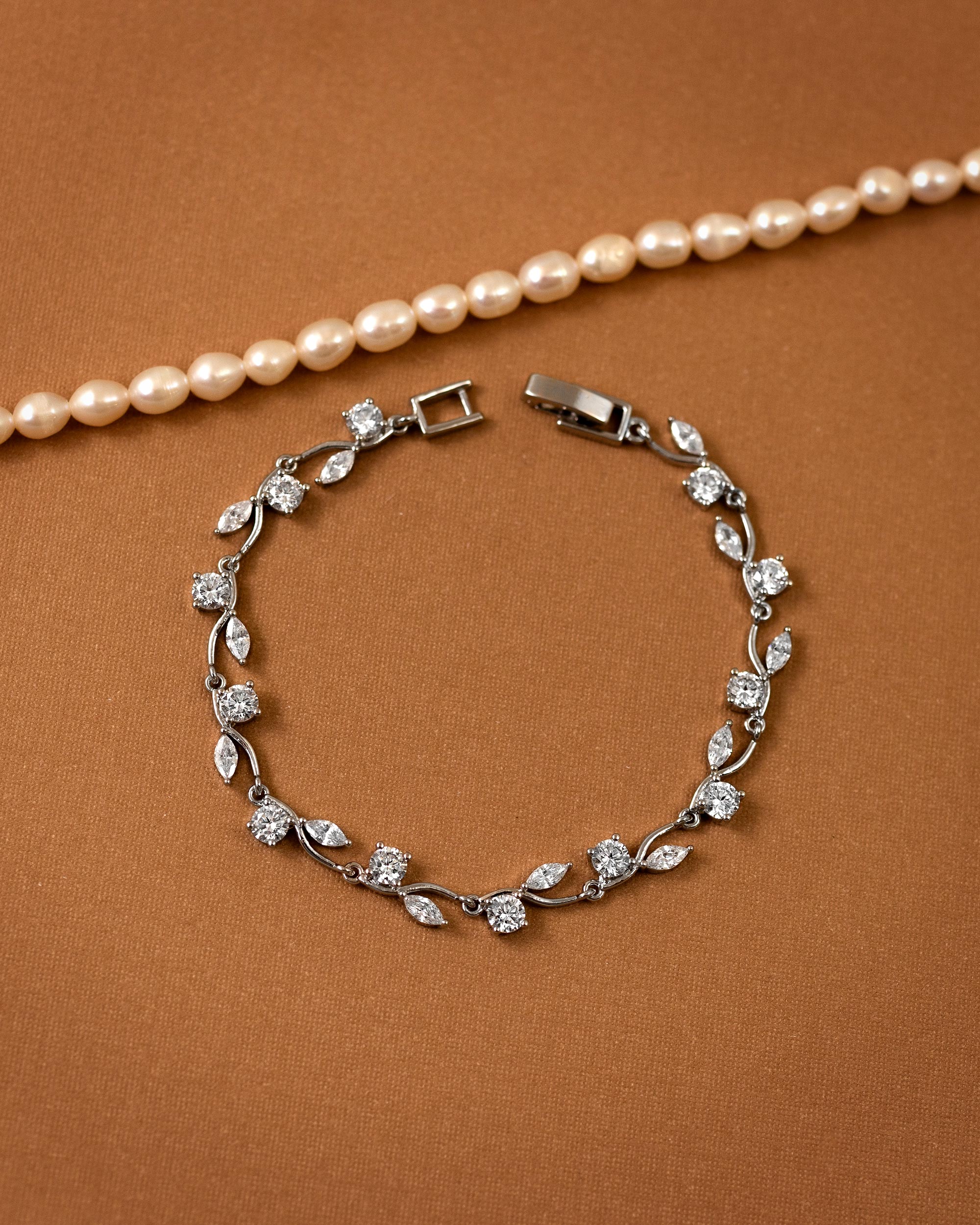 CZ Bracelet Marquise and Round Vine A silver-toned leaf and flower bracelet with clear stones, displayed on a warm brown textured background with a pearl necklace partially visible at the top.