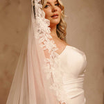 Cathedral Mantilla Wedding Veil Adorned with Lace Cassandra Lynne -A woman wearing a wedding dress and a long sheer tulle veil with floral lace applique, posed against a warm beige studio background.