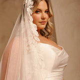 Cathedral Mantilla Wedding Veil Adorned with Lace Cassandra Lynne -A woman wearing a bridal veil with floral lace trim and a strapless wedding dress, posed against a warm beige background.