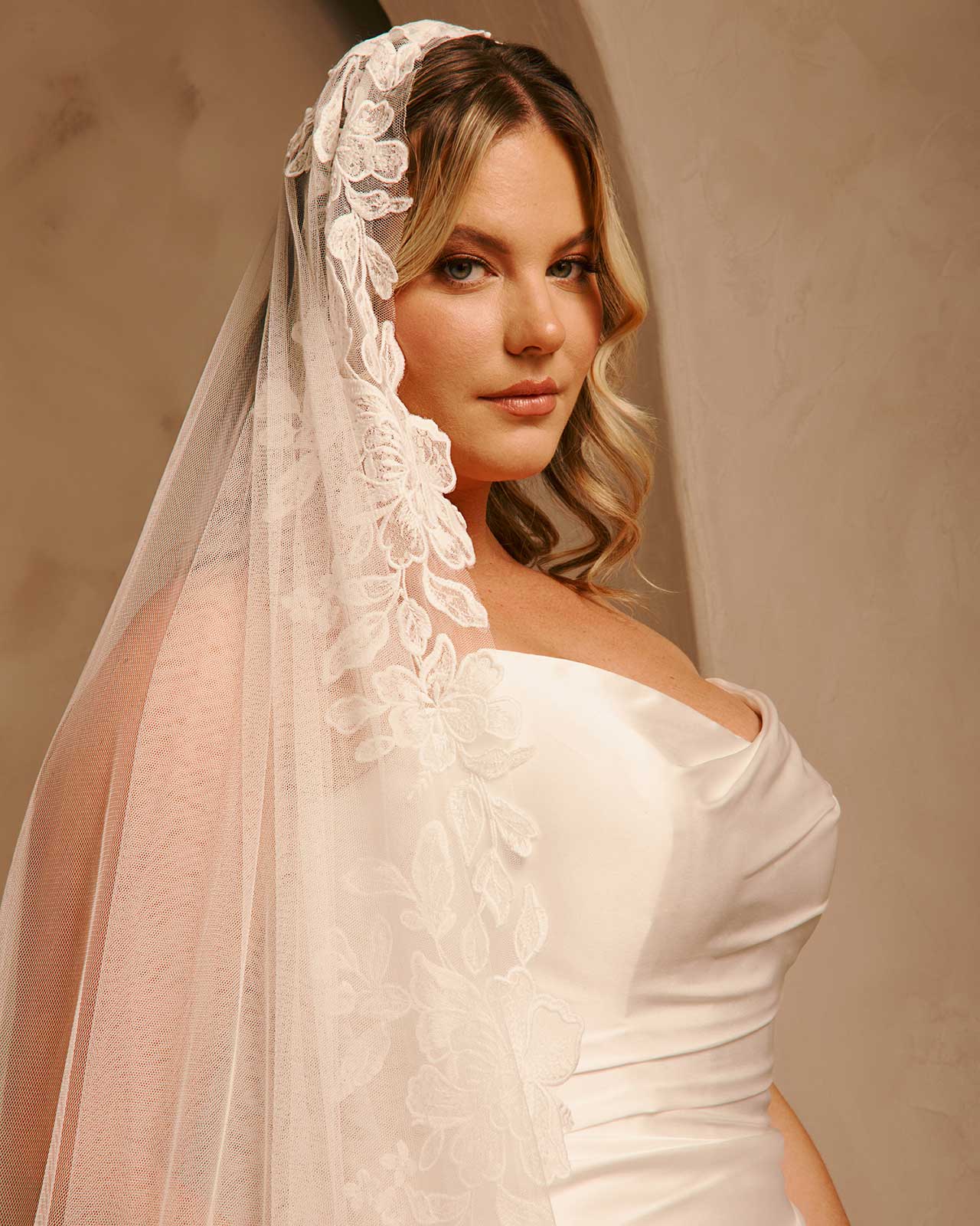 Cathedral Mantilla Wedding Veil Adorned with Lace Cassandra Lynne -A woman wearing a bridal veil with floral lace trim and a strapless wedding dress, posed against a warm beige background.