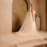 Cathedral Mantilla Wedding Veil Adorned with Lace Cassandra Lynne - A woman wearing a strapless wedding gown with a long sheer veil stands on stone steps in a warm beige interior with arched alcoves and a large vase in the background.