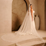 Cathedral Mantilla Wedding Veil Adorned with Lace Cassandra Lynne - A woman wearing a strapless wedding gown with a long sheer veil stands on stone steps in a warm beige interior with arched alcoves and a large vase in the background.