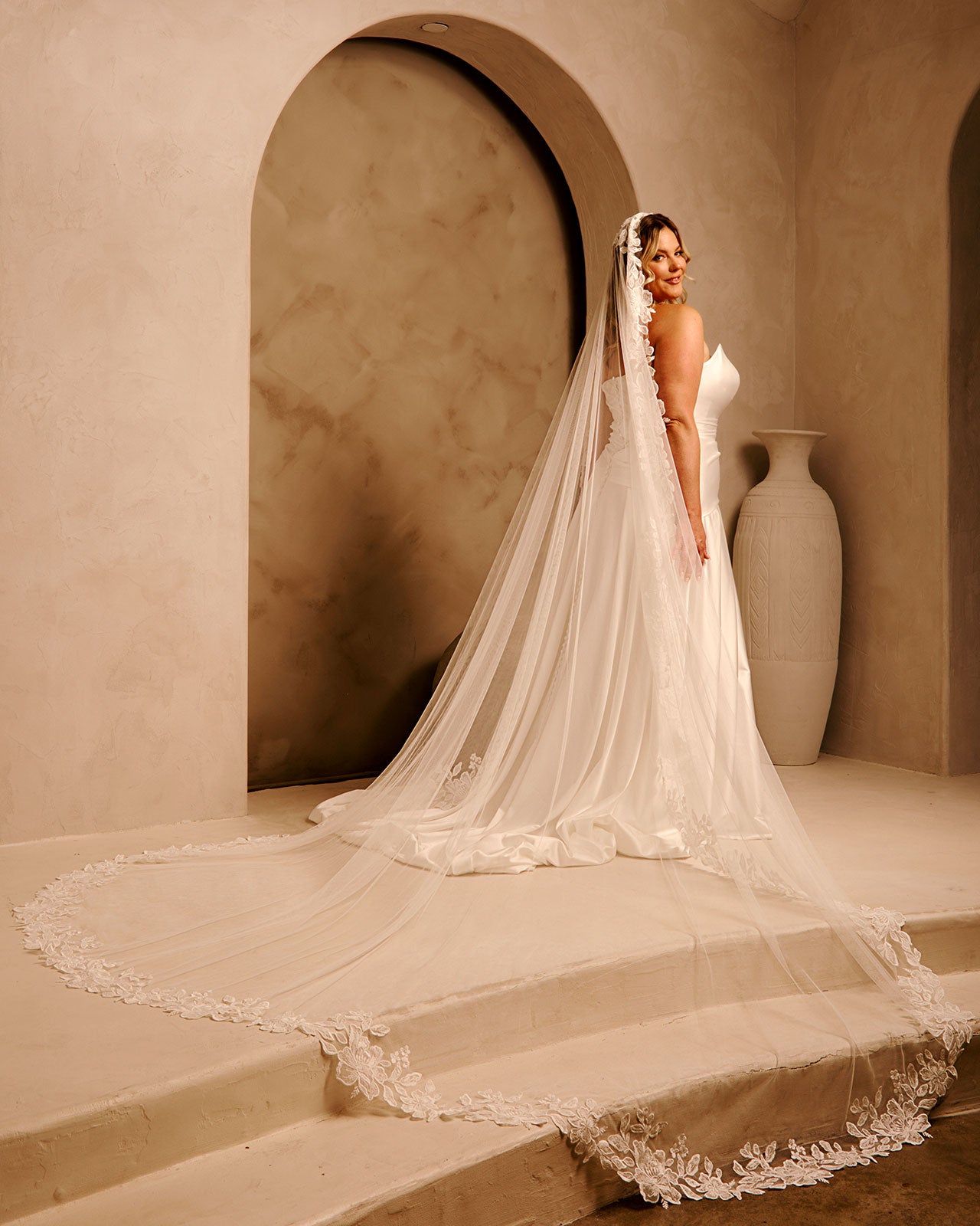 Cathedral Mantilla Wedding Veil Adorned with Lace Cassandra Lynne - A woman wearing a strapless wedding gown with a long sheer veil stands on stone steps in a warm beige interior with arched alcoves and a large vase in the background.
