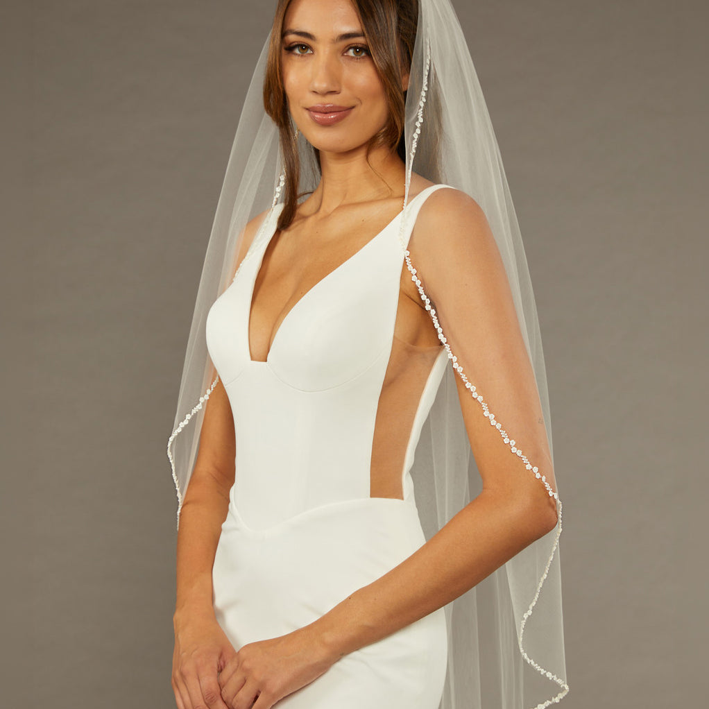 Metal Flower and Beads Fingertip Bridal Veil Woman wearing a  wedding dress with a sheer veil against a brown background