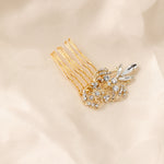 Dainty Regal Wedding Hair Comb A gold hair comb accessory with a floral and leaf design embellished with clear rhinestones, resting on a soft beige fabric background.