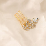 Dainty Regal Wedding Hair Comb A gold hair comb accessory with a floral and leaf design embellished with clear rhinestones, resting on a soft beige fabric background.