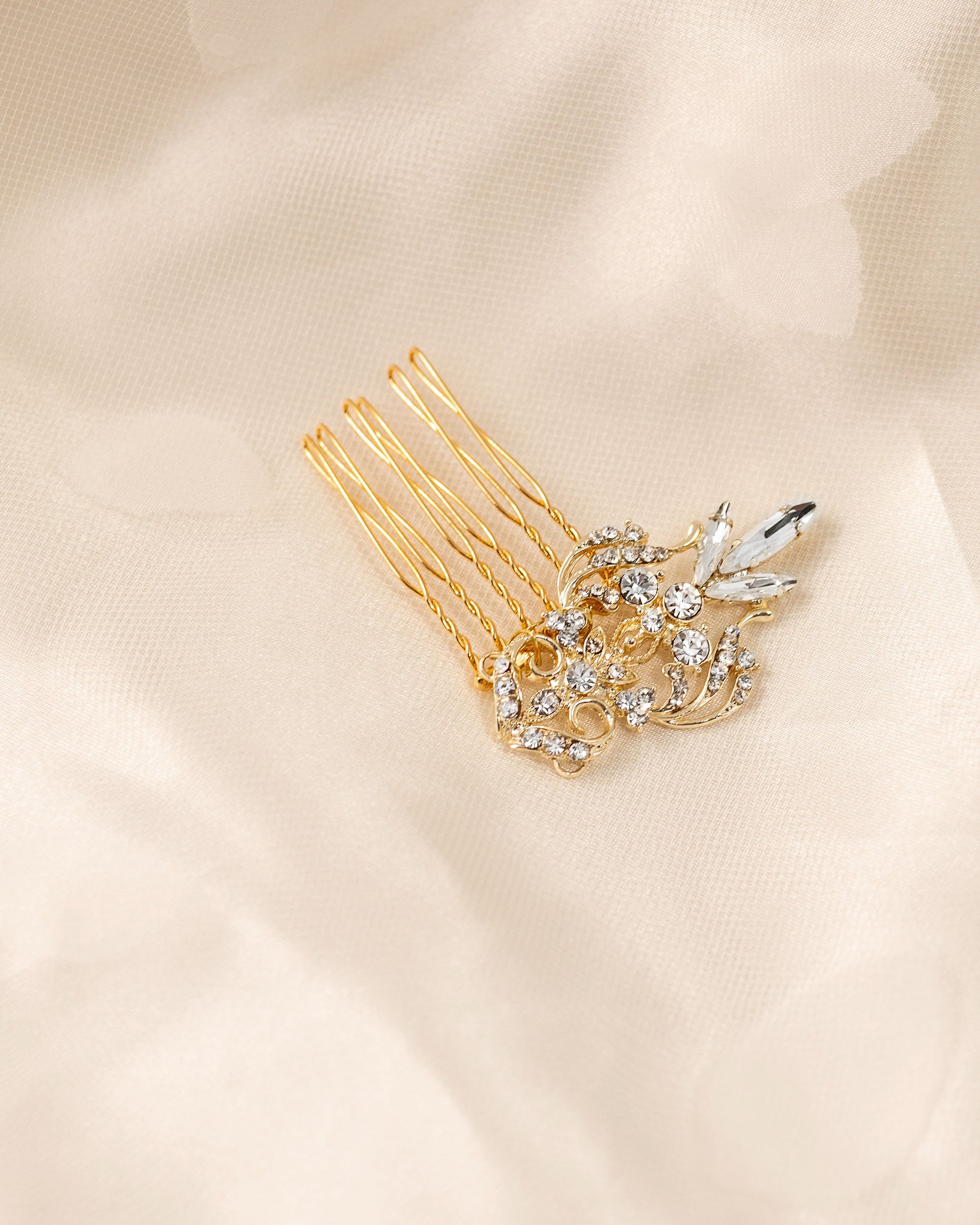 Dainty Regal Wedding Hair Comb A gold hair comb accessory with a floral and leaf design embellished with clear rhinestones, resting on a soft beige fabric background.