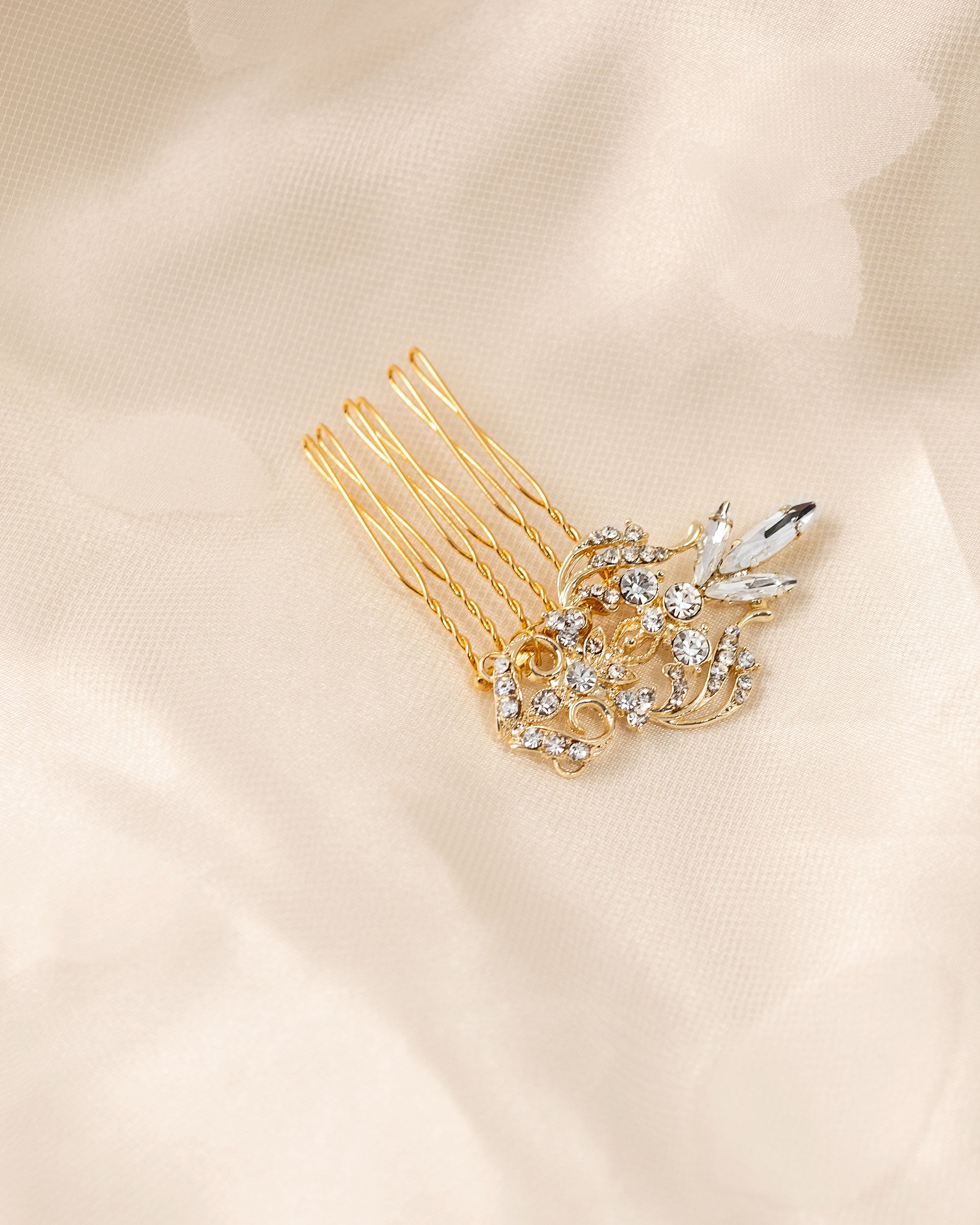 Dainty Regal Wedding Hair Comb A gold hair comb accessory with a floral and leaf design embellished with clear rhinestones, resting on a soft beige fabric background.