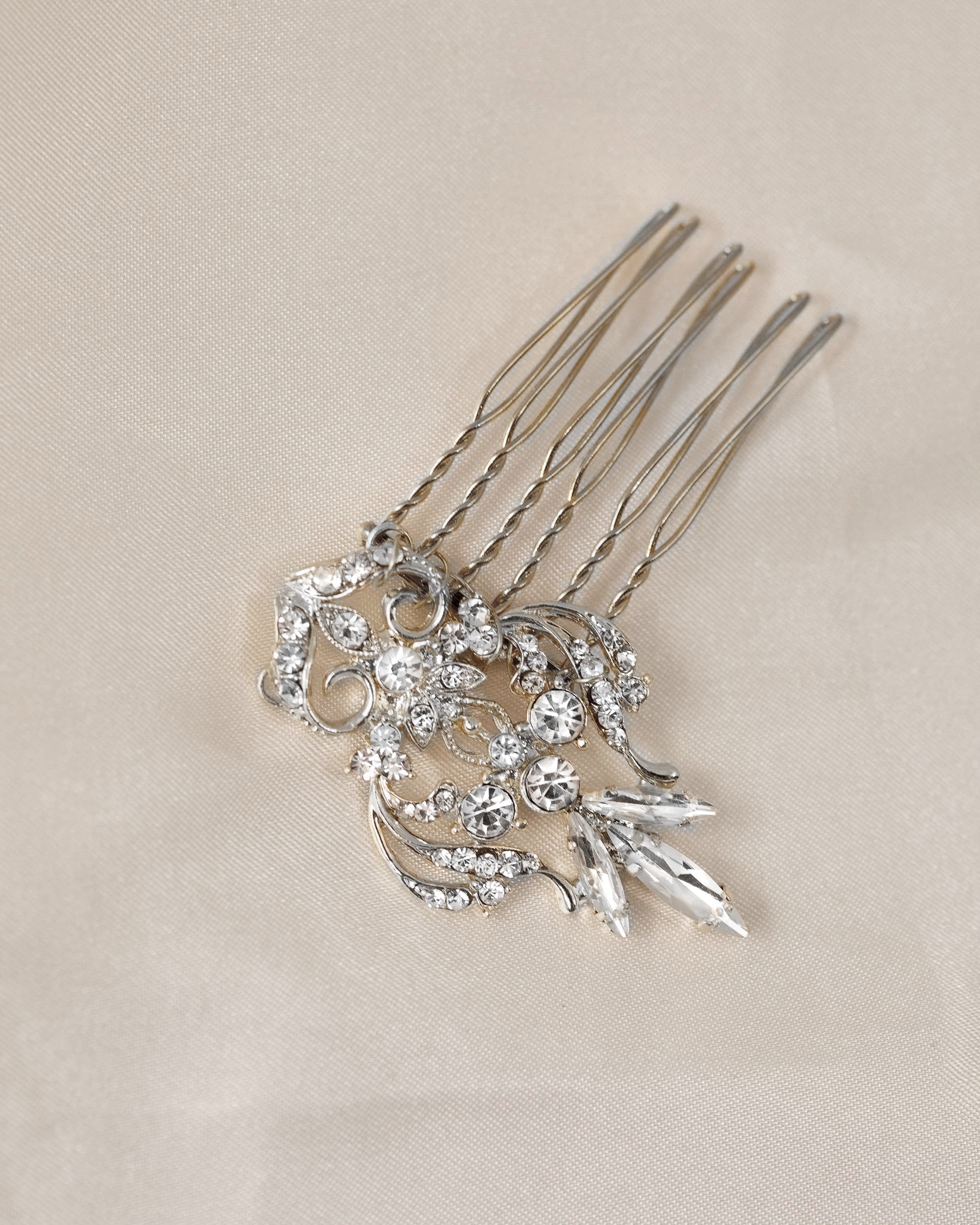Dainty Regal Wedding Hair Comb A silver-toned hair comb accessory with ornate floral and leaf designs set with clear rhinestones, displayed on a textured beige fabric background.
