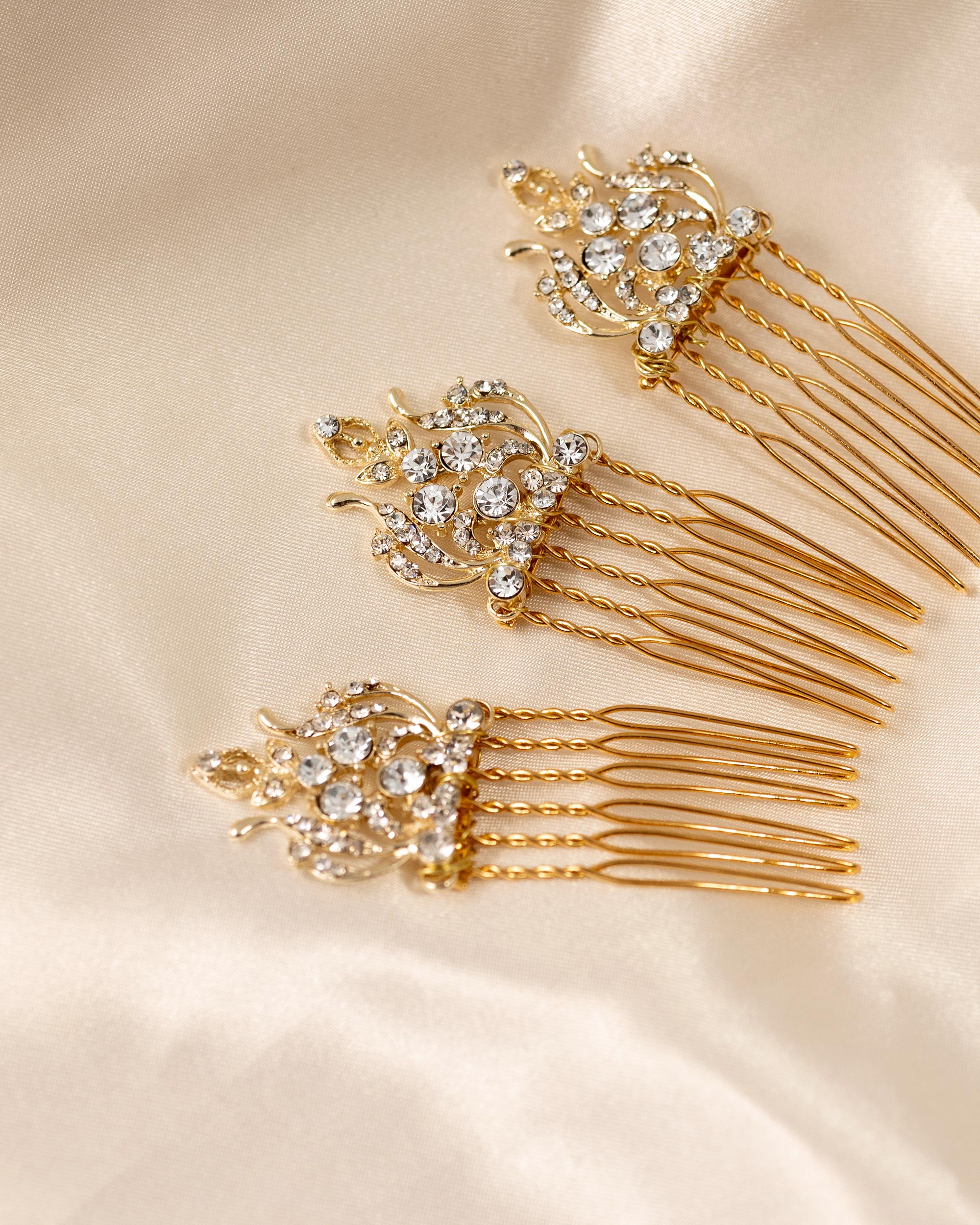 Regal Styled Small Wedding Hair Comb Three gold bridal hair combs embellished with clear rhinestones, arranged diagonally on a cream satin fabric background.