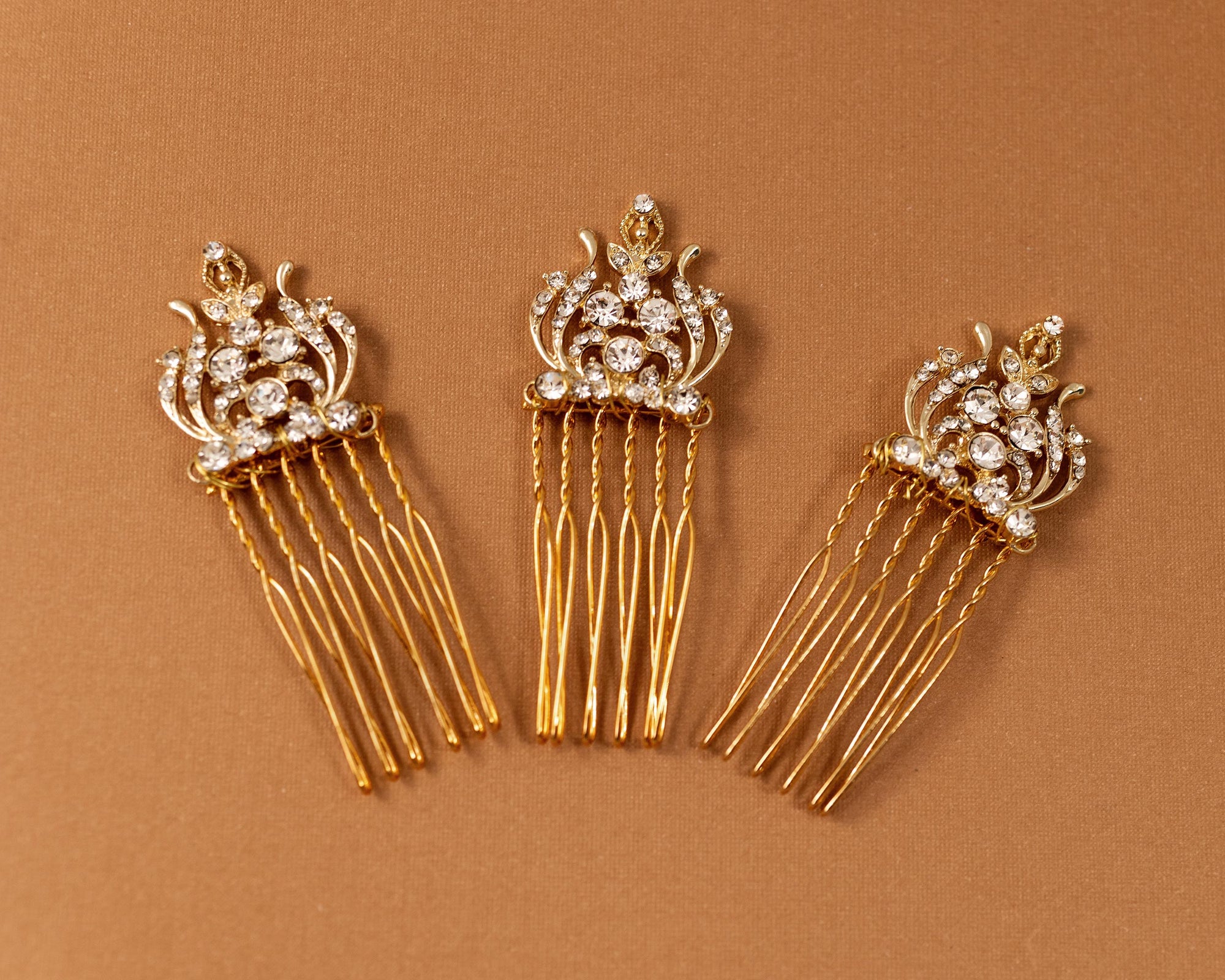 Regal Styled Small Wedding Hair Comb Three gold hair combs with ornate crystal-embellished crowns and long dangling strands, arranged on a warm brown background.