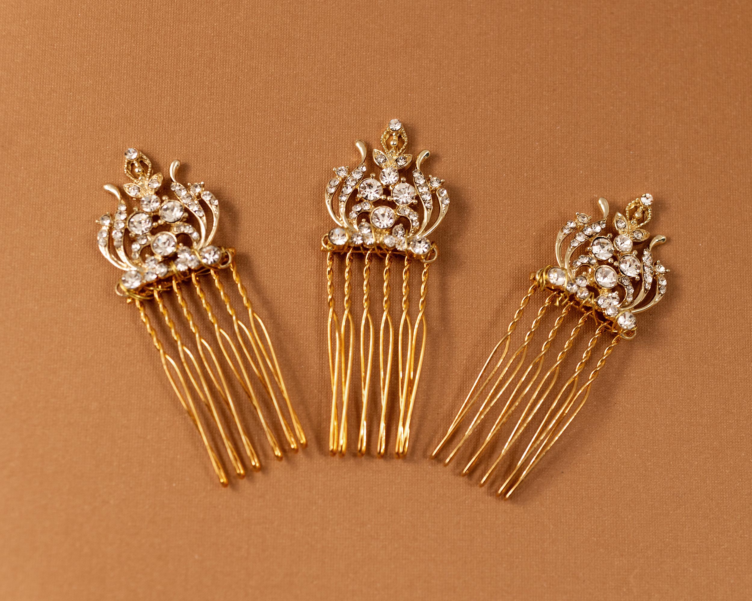 Regal Styled Small Wedding Hair Comb Three gold hair combs with ornate crystal-embellished crowns and long dangling strands, arranged on a warm brown background.