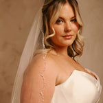 Cathedral Veil with Oat Pearl Scallop Edge A woman wearing a white strapless wedding dress and sheer veil, with a delicate beaded vine draped down her shoulder, posed against a warm beige studio backdrop.
