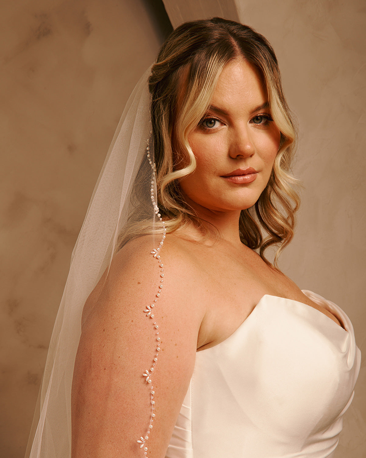 Cathedral Veil with Oat Pearl Scallop Edge A woman wearing a white strapless wedding dress and sheer veil, with a delicate beaded vine draped down her shoulder, posed against a warm beige studio backdrop.