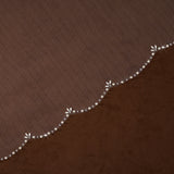 Cathedral Veil with Oat Pearl Scallop Edge A close-up of a brown textured fabric with a decorative wavy border featuring small white floral motifs and clear rhinestone accents.