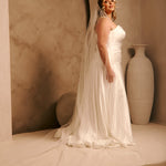 Cathedral Veil with Oat Pearl Scallop Edge A woman wearing a sleeveless ivory wedding gown with a long sheer veil stands in a studio setting with warm beige walls, an arched backdrop, and a large cream vase.