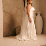 Cathedral Veil with Oat Pearl Scallop Edge A woman wearing a sleeveless ivory wedding gown with a long sheer veil stands in a studio setting with warm beige walls, an arched backdrop, and a large cream vase.