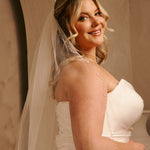 Cathedral Veil with Oat Pearl Scallop Edge A smiling blonde woman in a ivory strapless wedding dress with a sheer veil, photographed indoors against a warm beige wall.