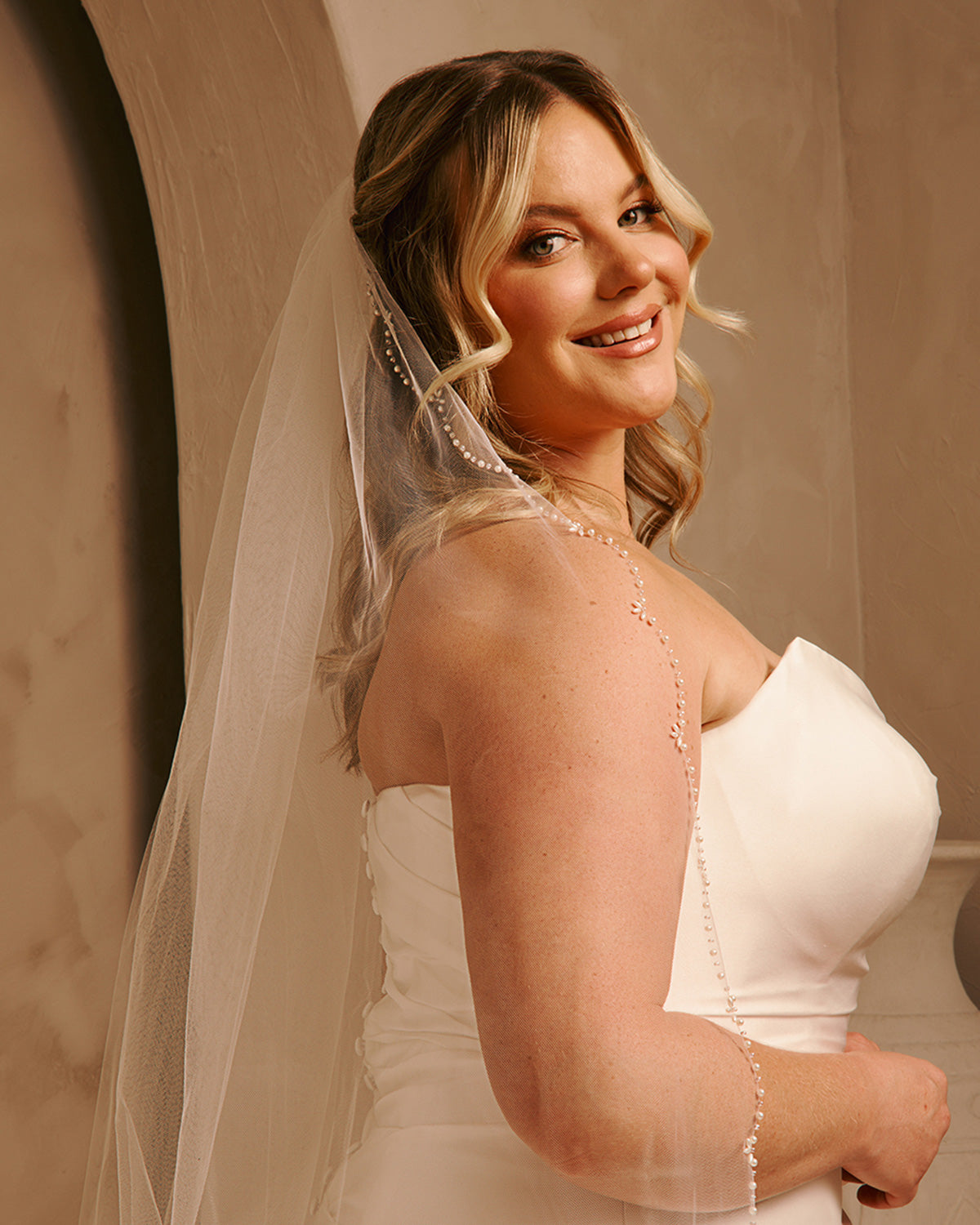 Cathedral Veil with Oat Pearl Scallop Edge A smiling blonde woman in a ivory strapless wedding dress with a sheer veil, photographed indoors against a warm beige wall.