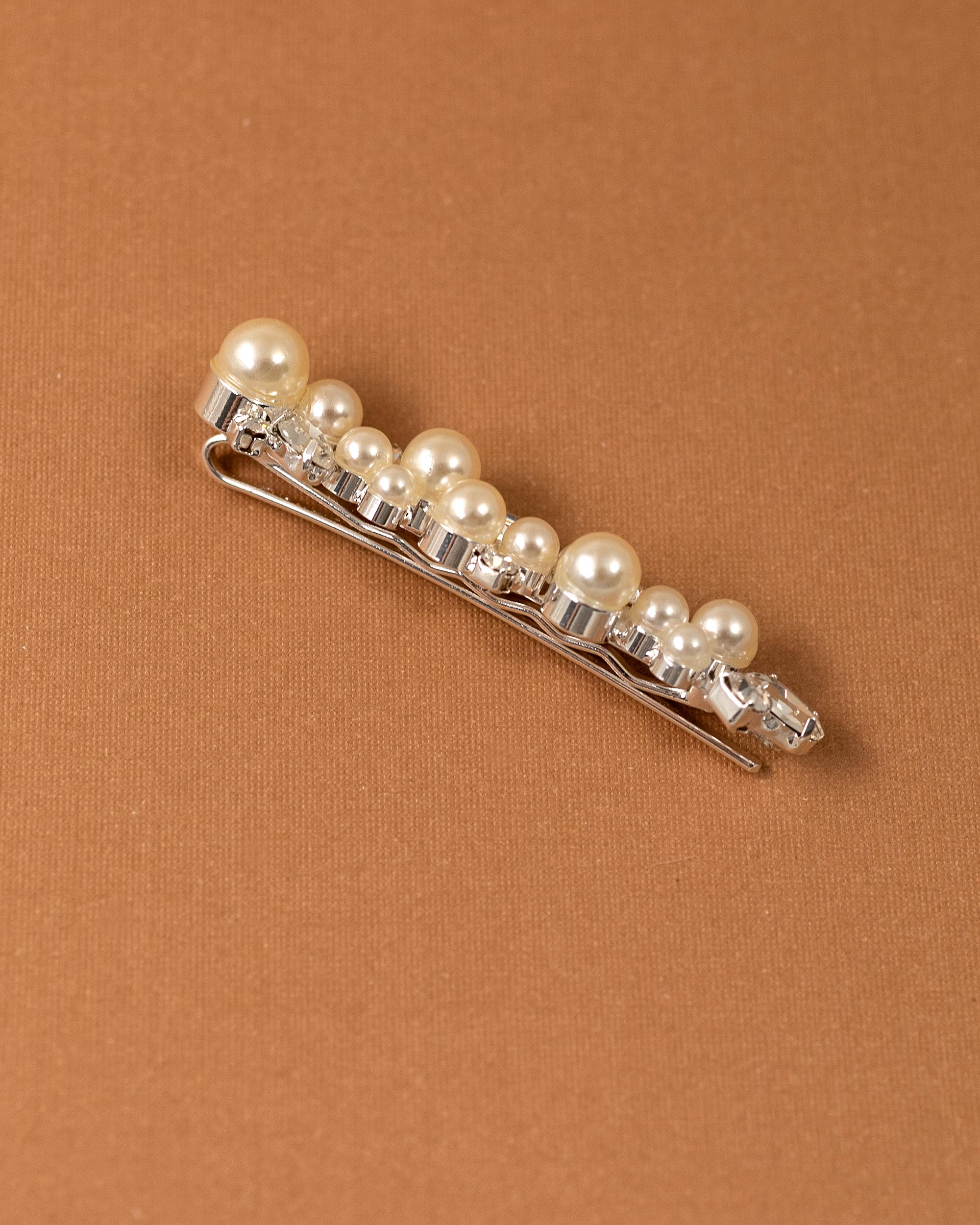 Bridal Bobby Pin with Ivory Pearls A silver hair claw clip embellished with a row of cream-colored faux pearls and clear rhinestones, displayed diagonally on a textured brown background.
