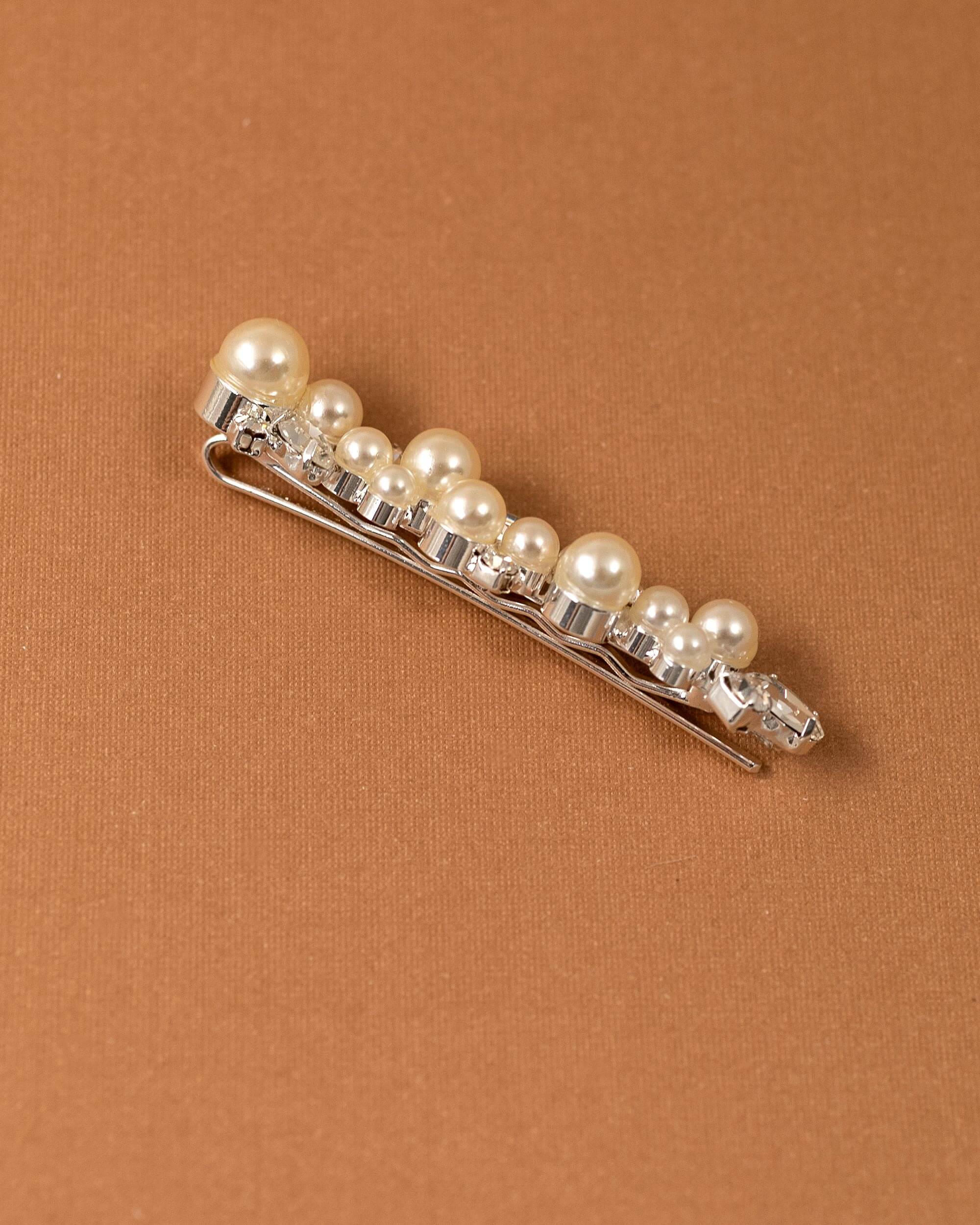 Bridal Bobby Pin with Ivory Pearls A silver hair claw clip embellished with a row of cream-colored faux pearls and clear rhinestones, displayed diagonally on a textured brown background.
