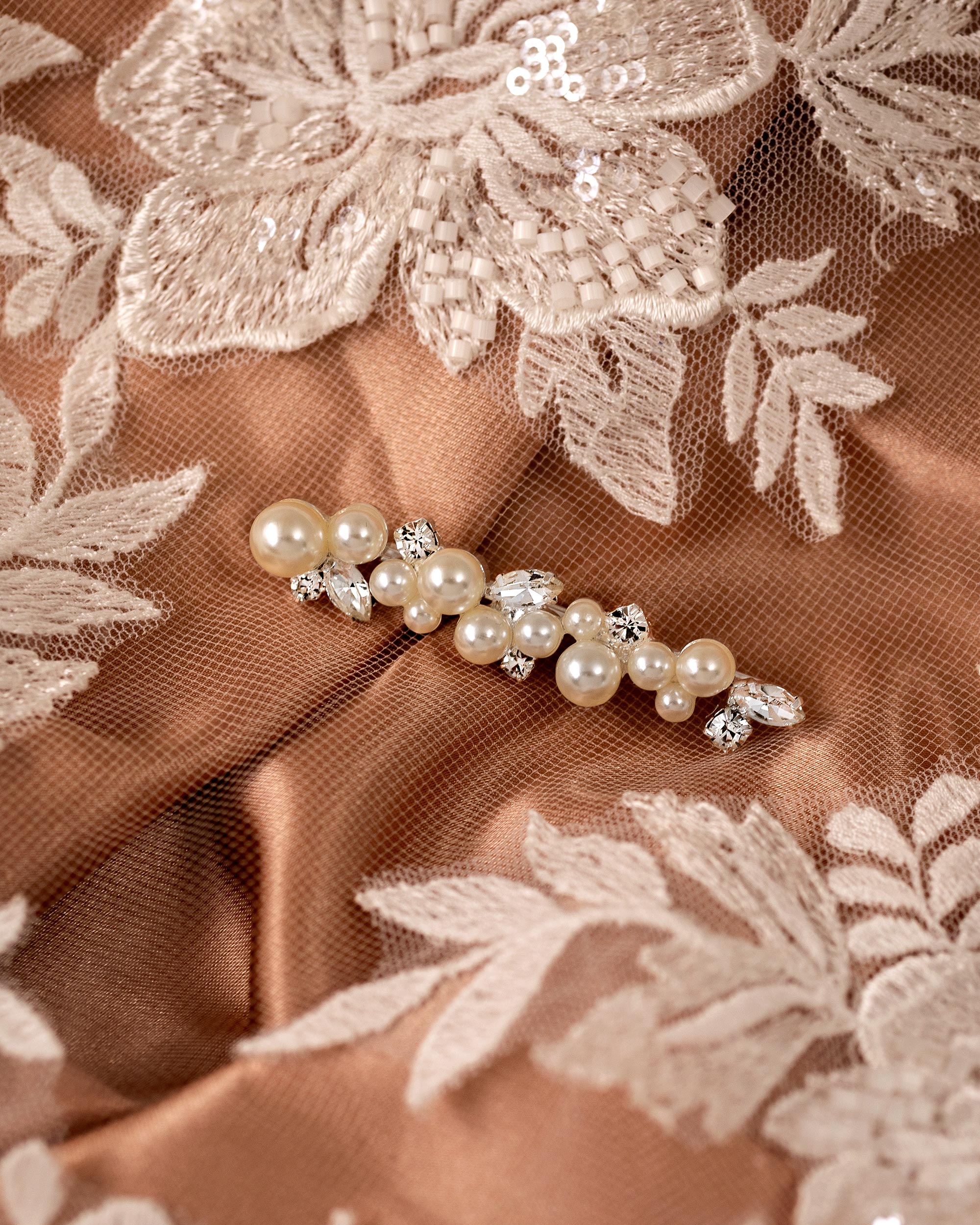 Bridal Bobby Pin with Ivory Pearls A close-up of a pearl and crystal hair accessory laid on warm taupe satin fabric with white floral lace embroidery.
