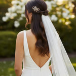 Pearl Swirl Rhinestone Wedding Hair Comb A woman seen from behind wearing a white wedding dress with an open back and a long sheer veil, with a decorative hair accessory in her hair, set against a soft-focus outdoor garden background.