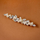 Bridal Bobby Pin with Marquise Jewels & Pearls A floral bridal hair clip featuring faux pearls and clear rhinestones, displayed diagonally on a warm brown textured background.