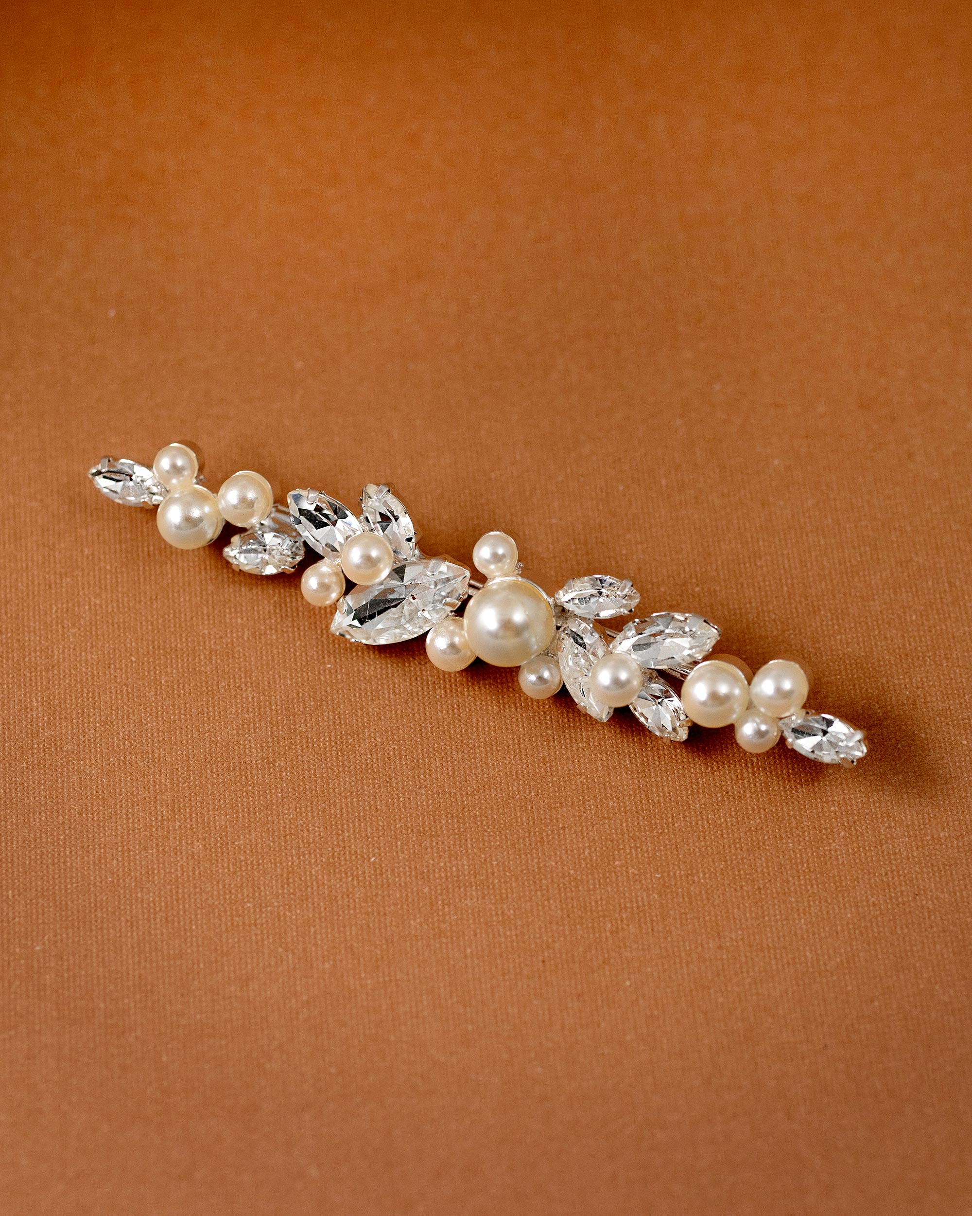 Bridal Bobby Pin with Marquise Jewels & Pearls A floral bridal hair clip featuring faux pearls and clear rhinestones, displayed diagonally on a warm brown textured background.