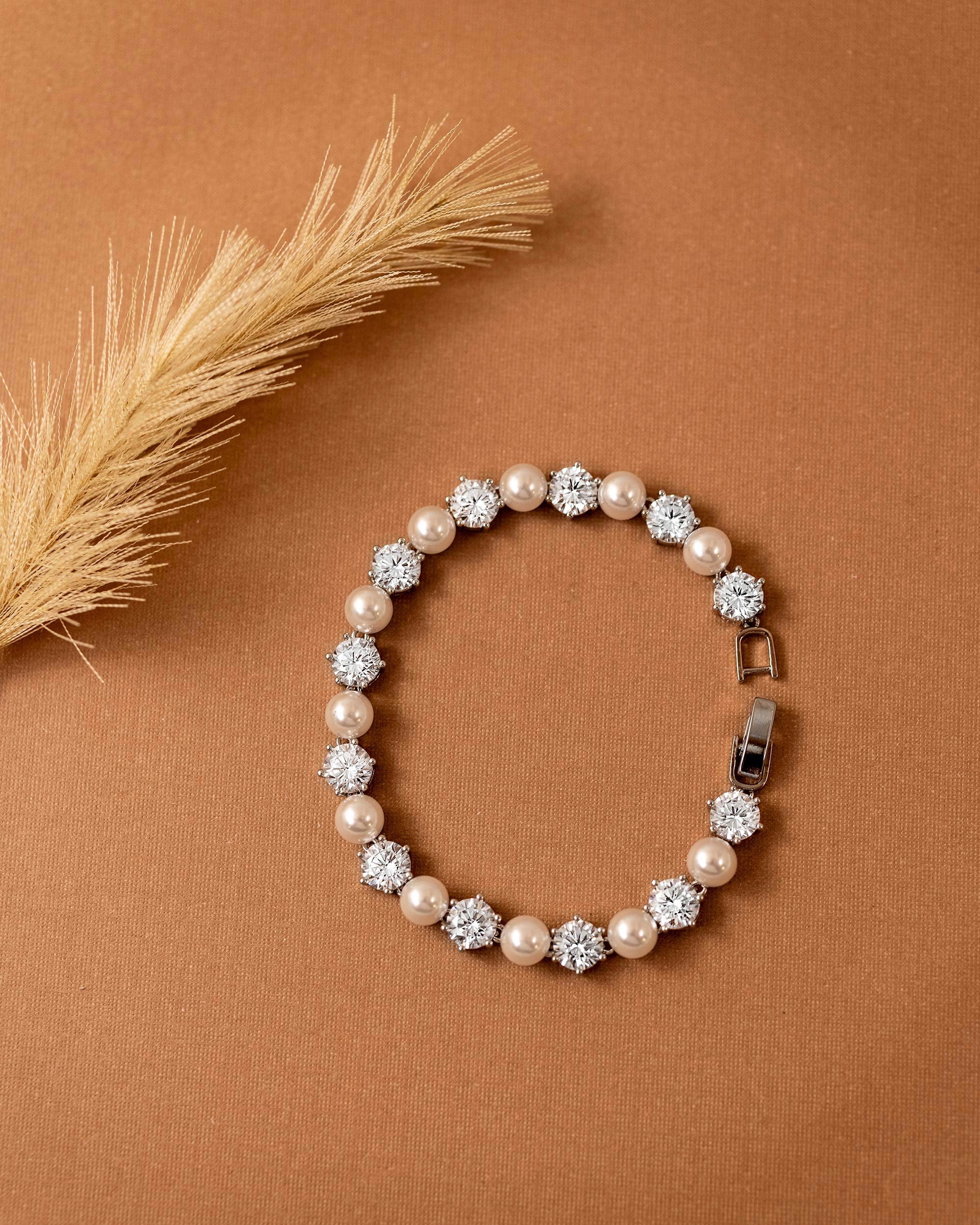 Pearl Bracelet Alternating CZ Stones A pearl and crystal beaded bracelet arranged in a circle on a warm brown textured background, with a dried grass stem in the upper left corner.
